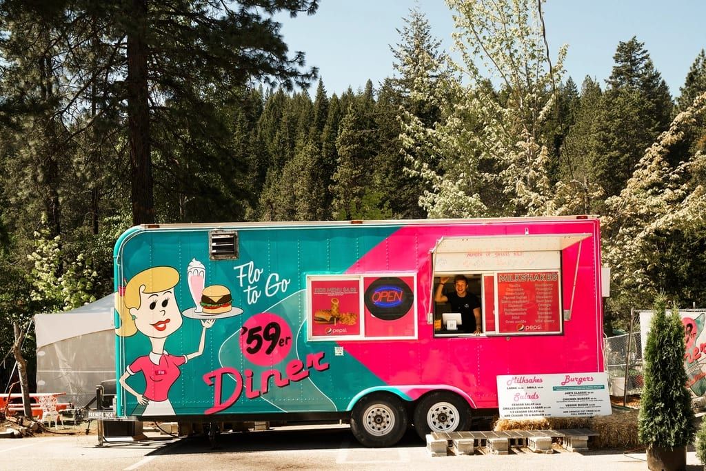 A retro pink and teal food trailer parked under pine trees